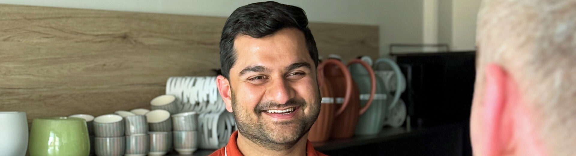 Mann in orange Polohemd lächelt und hält eine Tasse vor Regalen mit gestapeltem Geschirr, im Vordergrund unscharfe Schulter einer anderen Person.