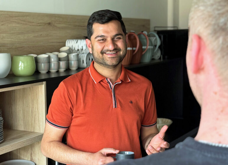 Mann in orange Polohemd lächelt und hält eine Tasse vor Regalen mit gestapeltem Geschirr, im Vordergrund unscharfe Schulter einer anderen Person.