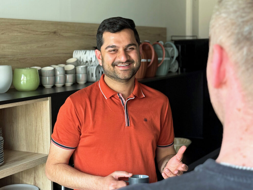 Mann in orange Polohemd lächelt und hält eine Tasse vor Regalen mit gestapeltem Geschirr, im Vordergrund unscharfe Schulter einer anderen Person.
