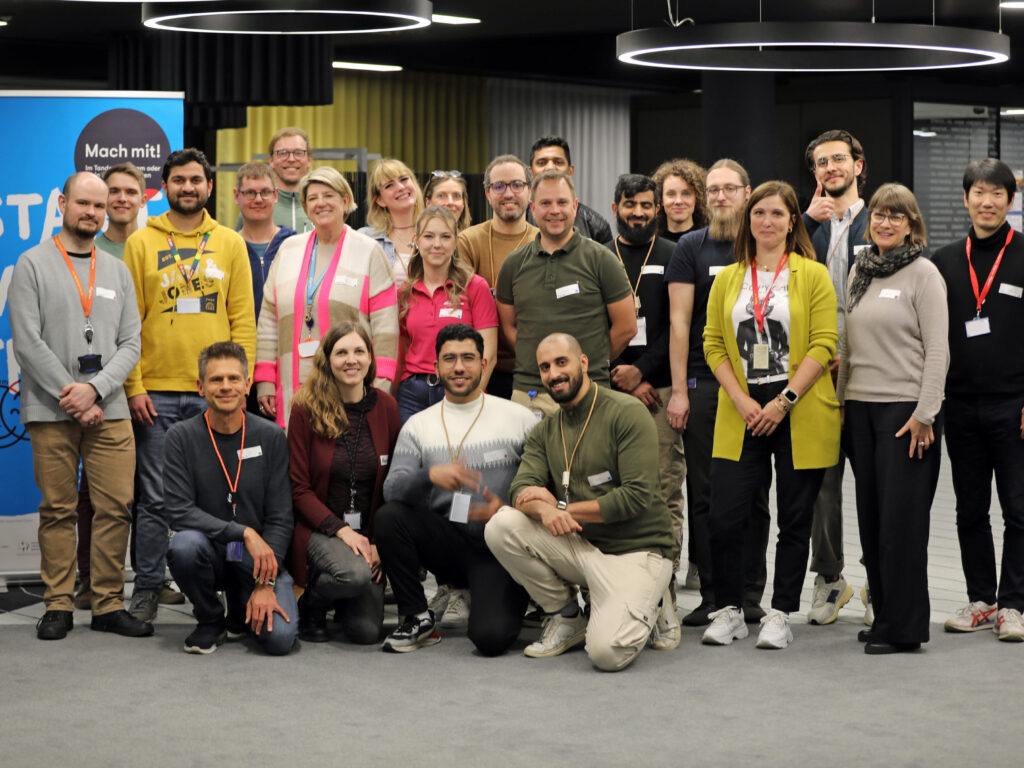 Gruppe von etwa 25 lächelnden Menschen verschiedener Altersgruppen in einem Innenraum vor einer Veranstaltungskulisse, einige kniend.