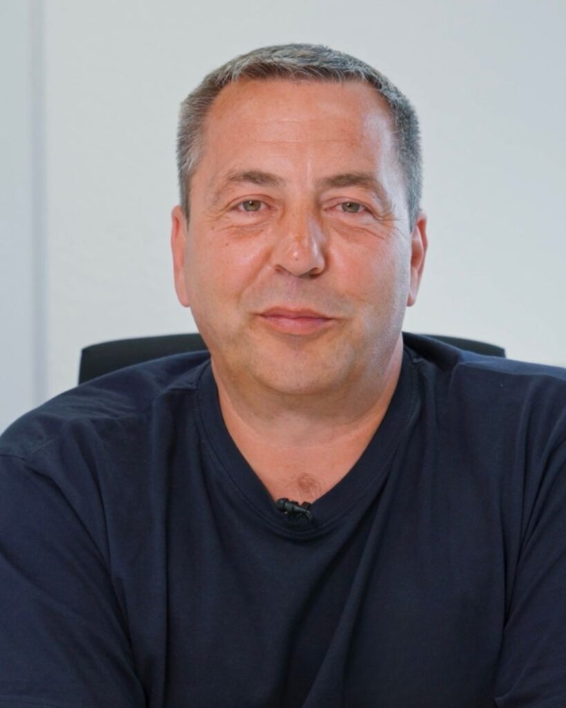 Mittelgroßer, lächelnder Mann mittleren Alters mit kurzem grauem Haar, dunkelblauem T‑Shirt und Mikrofon vor hellem Hintergrund.