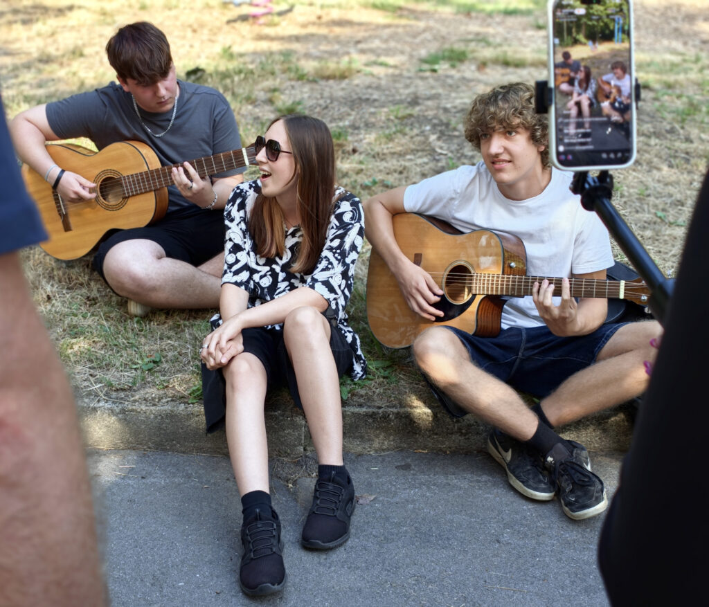 Drei Jugendliche sitzen im Gras, zwei spielen Gitarre, eine mit Sonnenbrille singt, daneben ein Smartphone auf einem Stativ filmt sie.