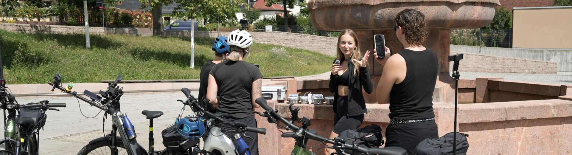 Vier Radfahrerinnen und Radfahrer vor einem steinernen Brunnen, eine Frau spricht, andere filmen oder hören zu, E‑Bikes im Vordergrund