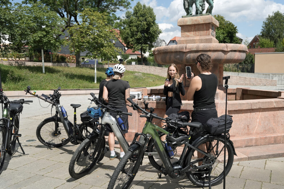 Vier Radfahrerinnen und Radfahrer vor einem steinernen Brunnen, eine Frau spricht, andere filmen oder hören zu, E‑Bikes im Vordergrund