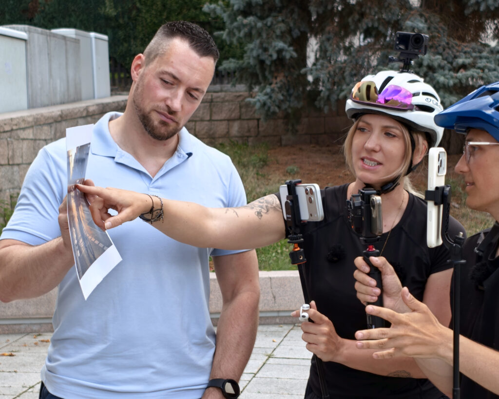 Zwei Radfahrer mit Helmen und Action‑Cams betrachten mit einem Mann in Polohemd eine Fotoausdrucke und diskutieren.