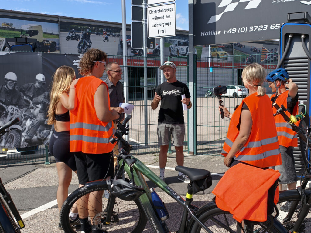 Gruppe von Fahrradfahrern in orangenen Warnwesten um einen Mann, der ihnen vor einem abgesperrten Rennstrecken-Eingang etwas erklärt.
