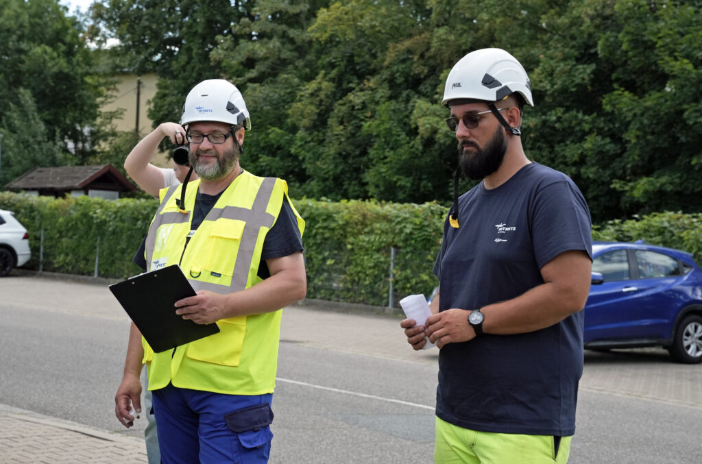 Zwei Arbeiter mit weißen Schutzhelmen, einer in gelber Warnweste mit Klemmbrett, der andere mit dunklem T‑Shirt und Handskizze in der Hand.