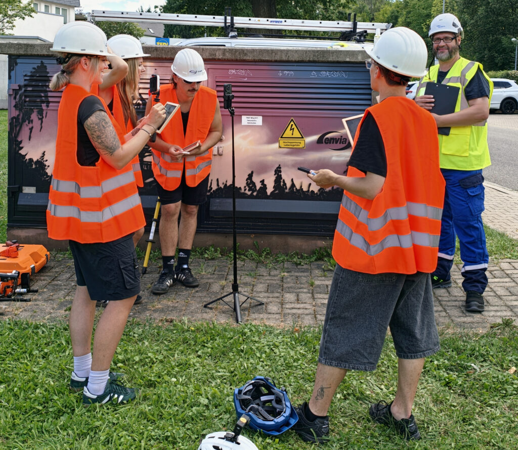 Fünf Personen in weißen Schutzhelmen und orangefarbigen Warnwesten stehen vor einem Stromkasten; eine Person hält Klemmbrett, andere Tablets und Kameras.