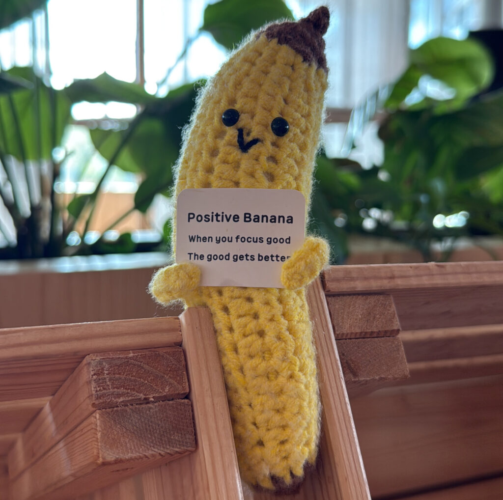 Gehäkelte gelbe Banane mit Knopfaugen und kleinem Schild „Positive Banana“ auf hellem Holz vor Pflanzenkulisse