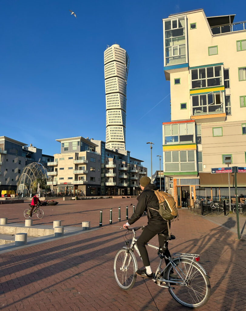 Radfahrer im Vordergrund vor markant verdrehtem Hochhaus (Turning Torso) und bunten Wohnhäusern an der Uferpromenade bei klarem Himmel.