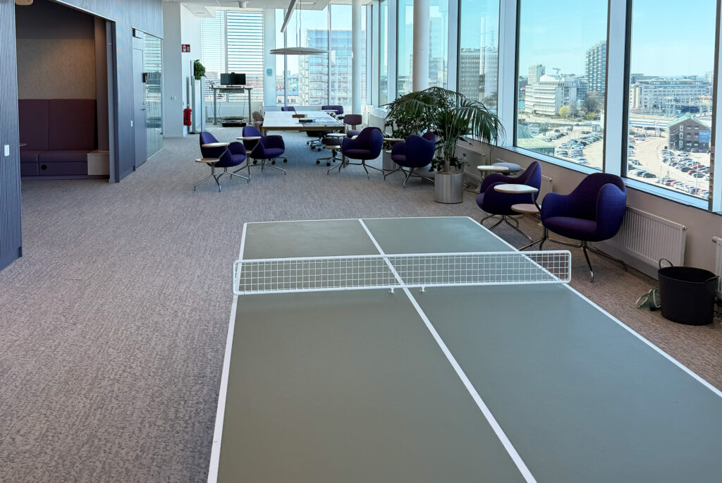 Büro-Lounge mit großem Fenster, violetten Sesseln, Pflanzen, Konferenztisch und einem Pingpong-Tisch in Vordergrund.
