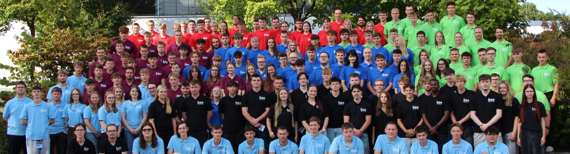 Großes Gruppenfoto vieler Jugendlicher und junger Erwachsener in farblich abgestuften Poloshirts (hellblau, dunkelblau, rot, grün, schwarz) vor Bäumen.