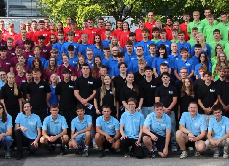 Großes Gruppenfoto vieler Jugendlicher und junger Erwachsener in farblich abgestuften Poloshirts (hellblau, dunkelblau, rot, grün, schwarz) vor Bäumen.
