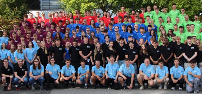 Großes Gruppenfoto vieler Jugendlicher und junger Erwachsener in farblich abgestuften Poloshirts (hellblau, dunkelblau, rot, grün, schwarz) vor Bäumen.