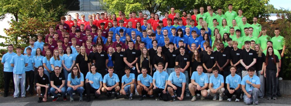 Großes Gruppenfoto vieler Jugendlicher und junger Erwachsener in farblich abgestuften Poloshirts (hellblau, dunkelblau, rot, grün, schwarz) vor Bäumen.