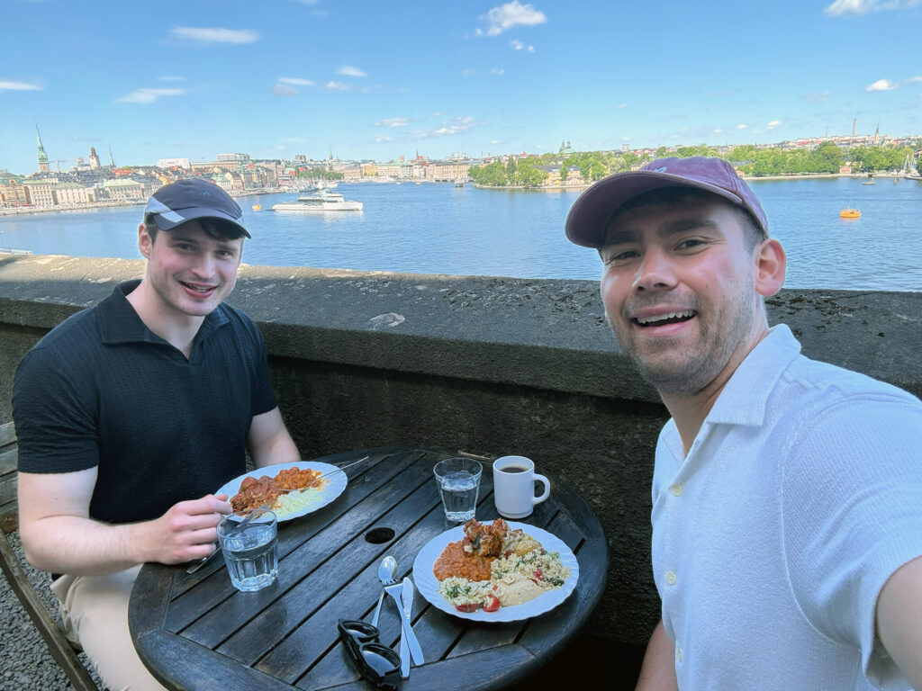 Zwei junge Männer mit Kappen essen auf einer Terrasse; im Hintergrund Blick über Wasser und Stadtsilhouette mit Booten.