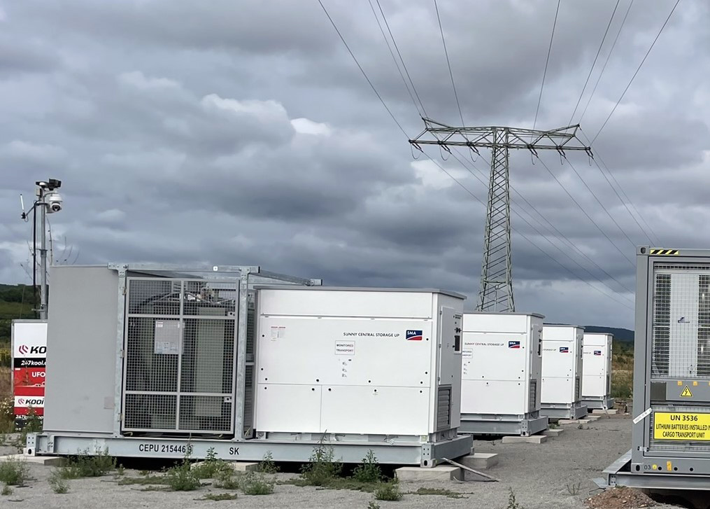 Reihe weißer Energiespeichercontainer auf Betonplatten unter wolkigem Himmel, im Hintergrund Hochspannungsmast und Überwachungskamera.
