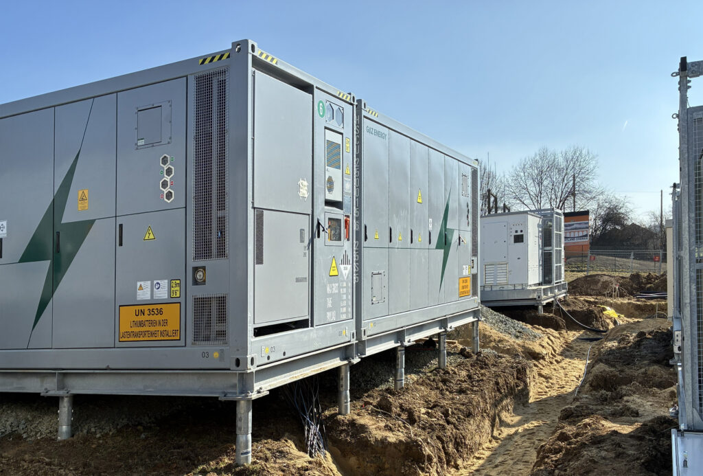 Große graue Energiespeichercontainer auf Stelzen neben ausgehobenen Kabelgräben auf einer Baustelle unter klarem Himmel.