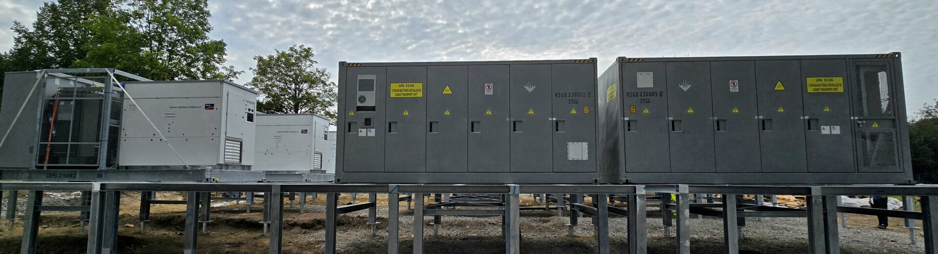 Reihenweise Industriecontainer auf Stahlstützen unter einem wolkenverhangenen Himmel, Bäume links im Hintergrund.