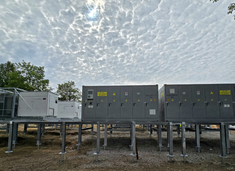 Reihenweise Industriecontainer auf Stahlstützen unter einem wolkenverhangenen Himmel, Bäume links im Hintergrund.