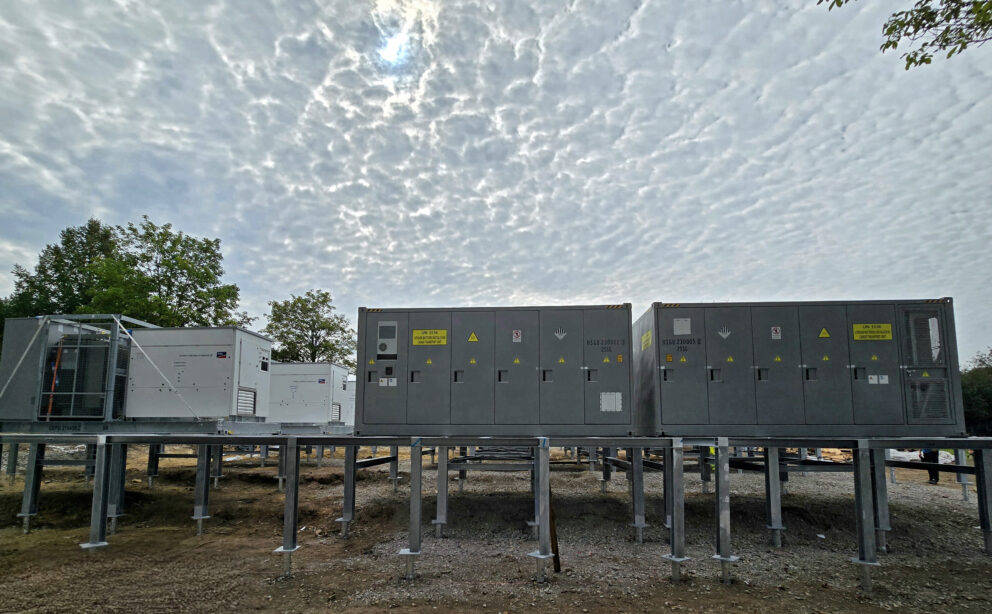Reihenweise Industriecontainer auf Stahlstützen unter einem wolkenverhangenen Himmel, Bäume links im Hintergrund.