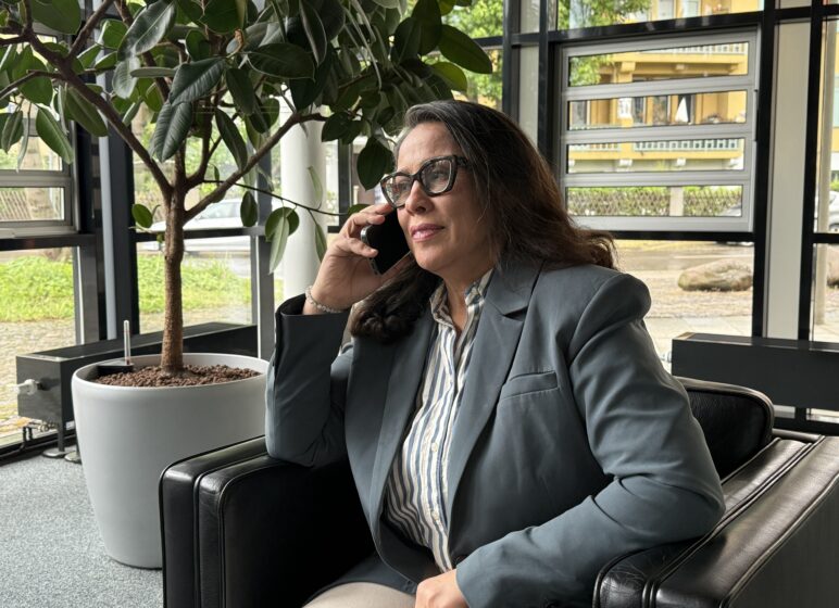 Frau mit Brille und grauem Blazer sitzt in einem Sessel und telefoniert, im Hintergrund große Topfpflanze und Fensterfront