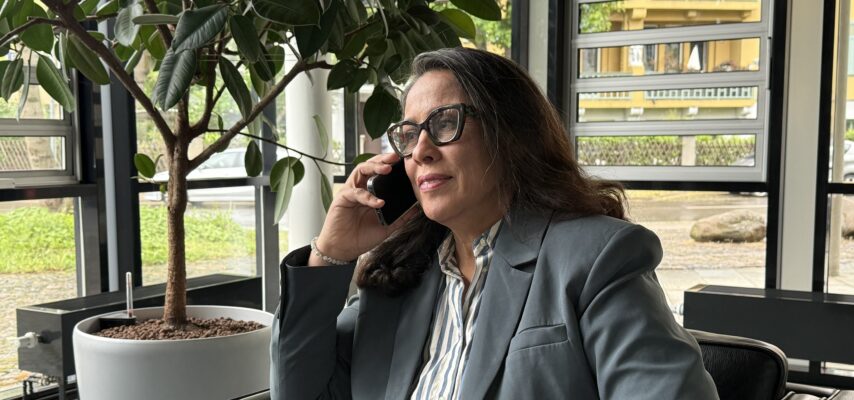 Frau mit Brille und grauem Blazer sitzt in einem Sessel und telefoniert, im Hintergrund große Topfpflanze und Fensterfront