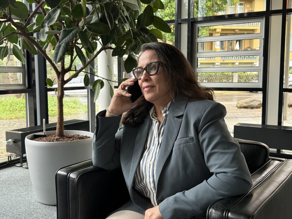 Frau mit Brille und grauem Blazer sitzt in einem Sessel und telefoniert, im Hintergrund große Topfpflanze und Fensterfront