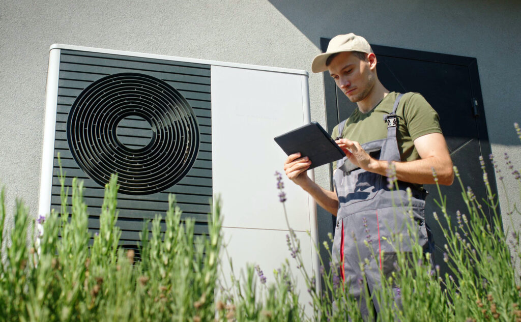 Installateur in Arbeitskleidung prüft mit Tablet eine Außeneinheit einer Wärmepumpe, Vordergrund mit Lavendelpflanzen.