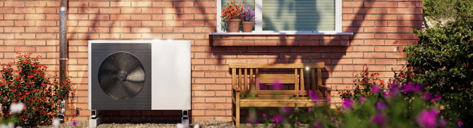 Außenwand eines Backsteinhauses mit Fenstersims, zwei Blumenkübeln, Holzbank und einer weißen Außeneinheit einer Wärmepumpe im Vorgarten.