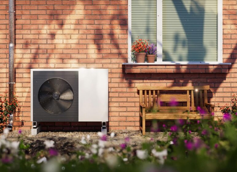 Außenwand eines Backsteinhauses mit Fenstersims, zwei Blumenkübeln, Holzbank und einer weißen Außeneinheit einer Wärmepumpe im Vorgarten.