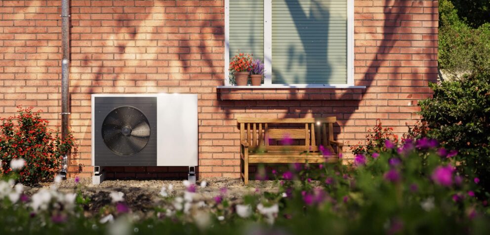 Außenwand eines Backsteinhauses mit Fenstersims, zwei Blumenkübeln, Holzbank und einer weißen Außeneinheit einer Wärmepumpe im Vorgarten.