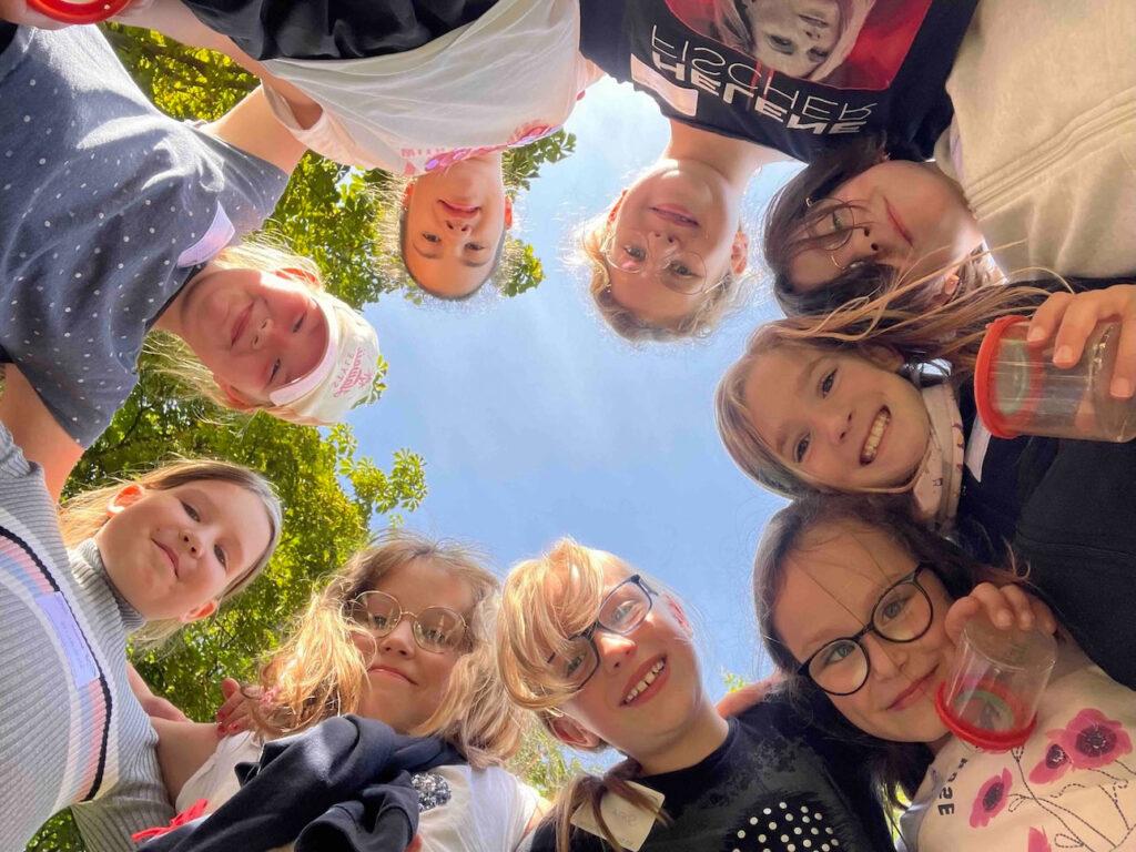 Neun lächelnde Kinder stehen im Kreis und blicken von oben in die Kamera, blauer Himmel und Baumkronen im Hintergrund.