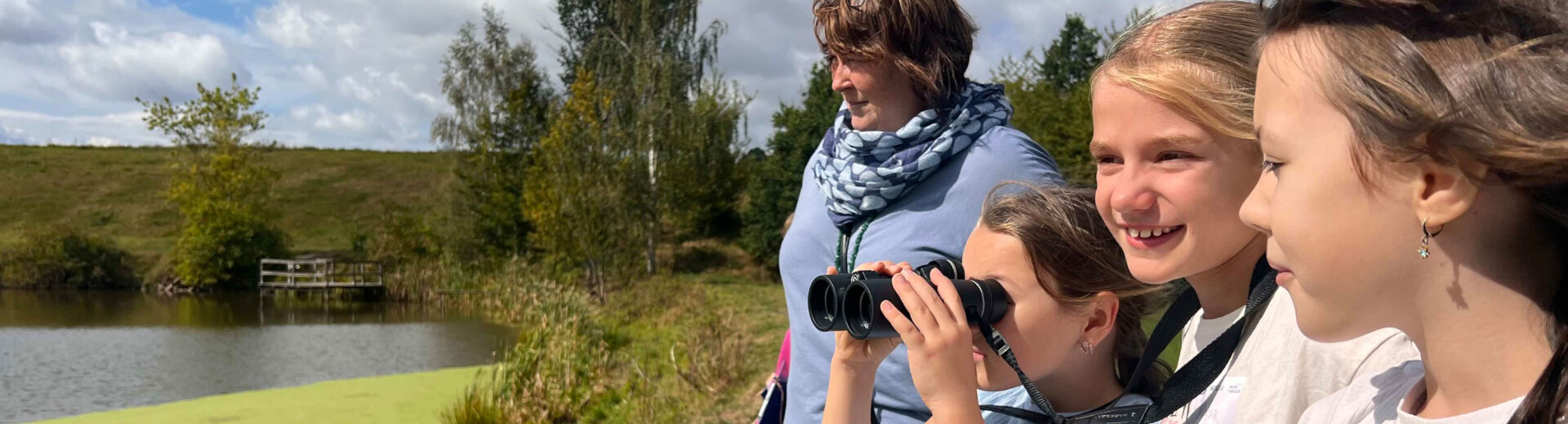 Vier lächelnde Kinder und eine Frau am Seeufer, ein Mädchen schaut durch Fernglas, grünes Wasser und Bäume im Hintergrund