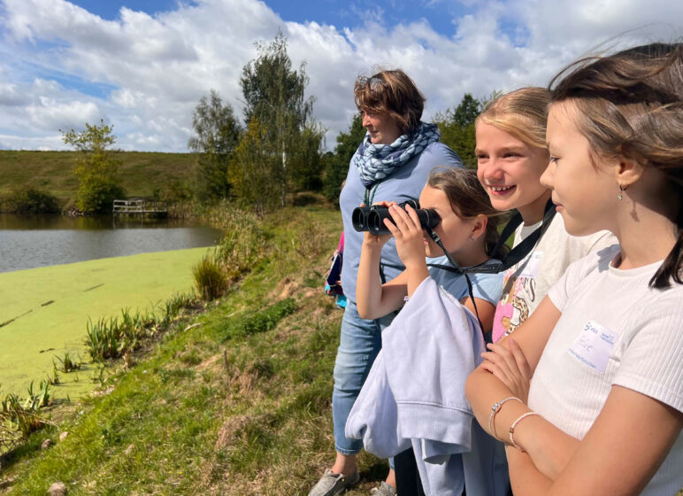 Vier lächelnde Kinder und eine Frau am Seeufer, ein Mädchen schaut durch Fernglas, grünes Wasser und Bäume im Hintergrund