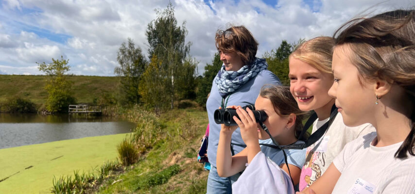 Vier lächelnde Kinder und eine Frau am Seeufer, ein Mädchen schaut durch Fernglas, grünes Wasser und Bäume im Hintergrund
