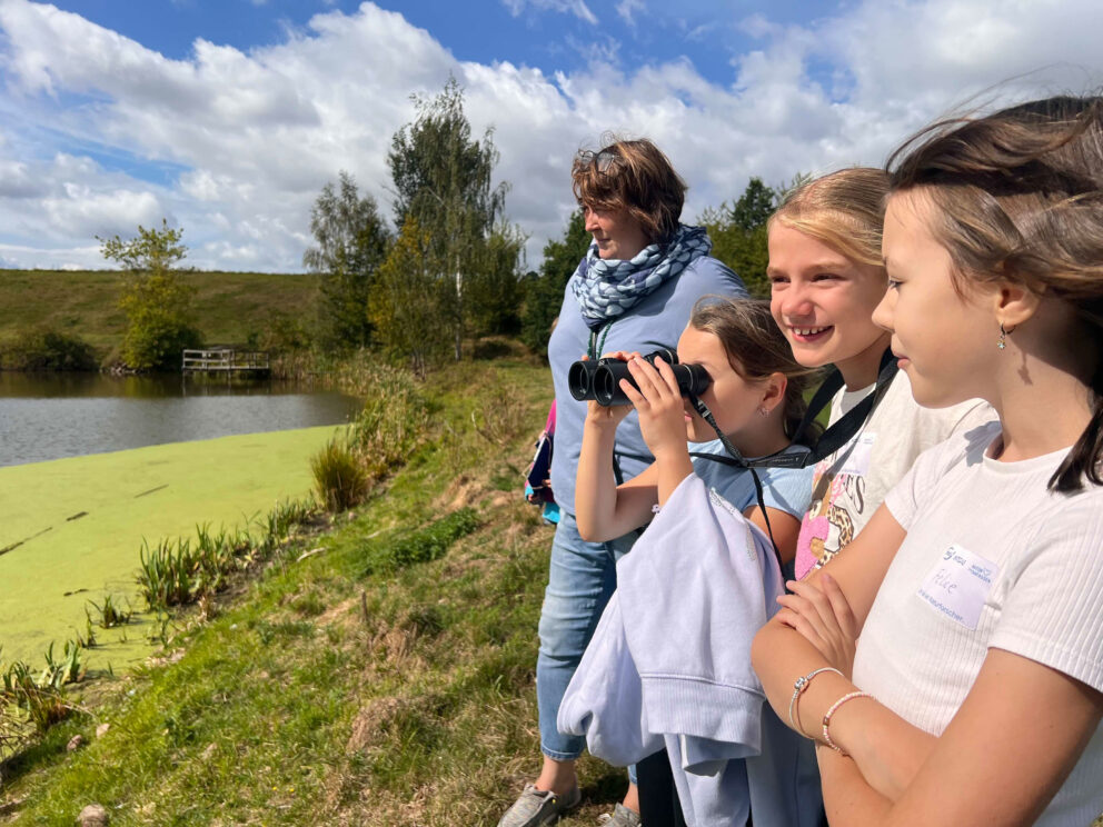 Vier lächelnde Kinder und eine Frau am Seeufer, ein Mädchen schaut durch Fernglas, grünes Wasser und Bäume im Hintergrund