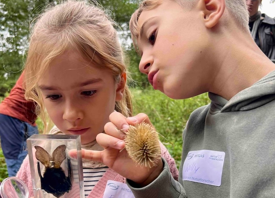 Zwei Kinder betrachten im Freien ein eingegossenes Insekt in einem Plexiglaswürfel; das Mädchen hält die Box, der Junge zeigt eine stachelige Samenkapsel.