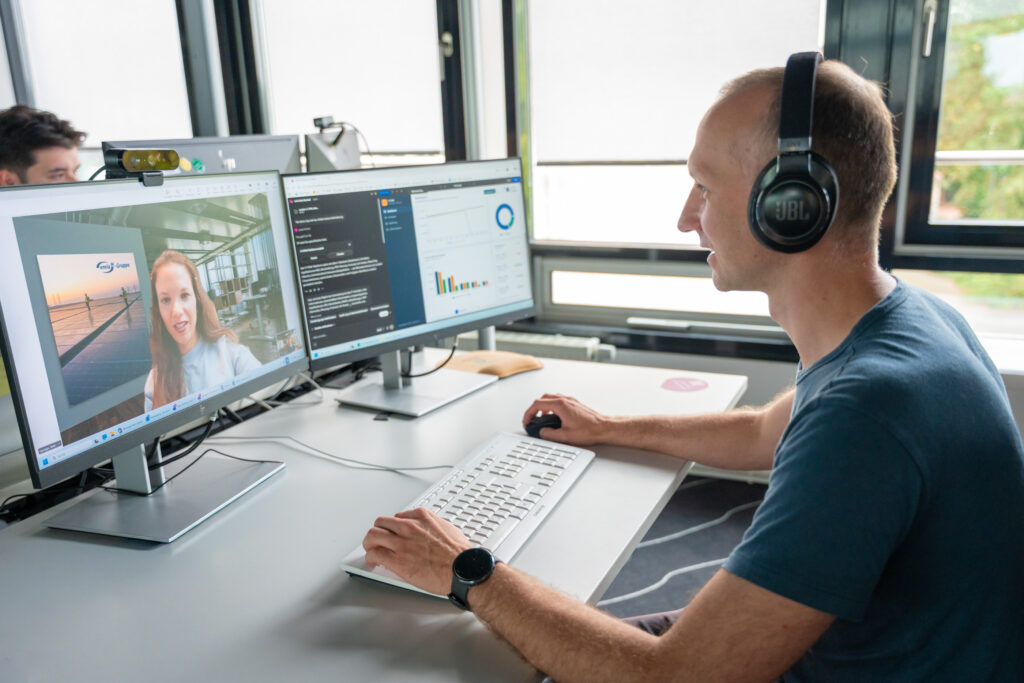Mann mit JBL-Kopfhörern arbeitet am Schreibtisch und führt Videokonferenz auf zwei Monitoren; links Gesicht einer Frau im Call.