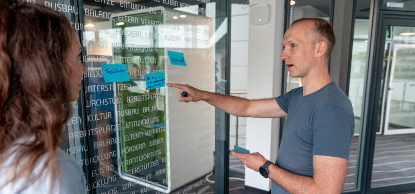 Zwei Mitarbeitende diskutieren vor einer Glaswand mit Klebezetteln; ein Mann zeigt mit dem Finger auf einen blauen Zettel.