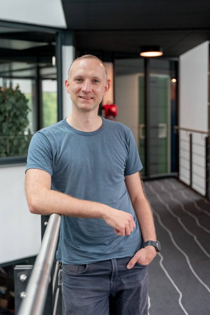 Männlicher Erwachsener in blauem T‑Shirt lehnt an einem Geländer in modernem Bürogebäude und lächelt in die Kamera.
