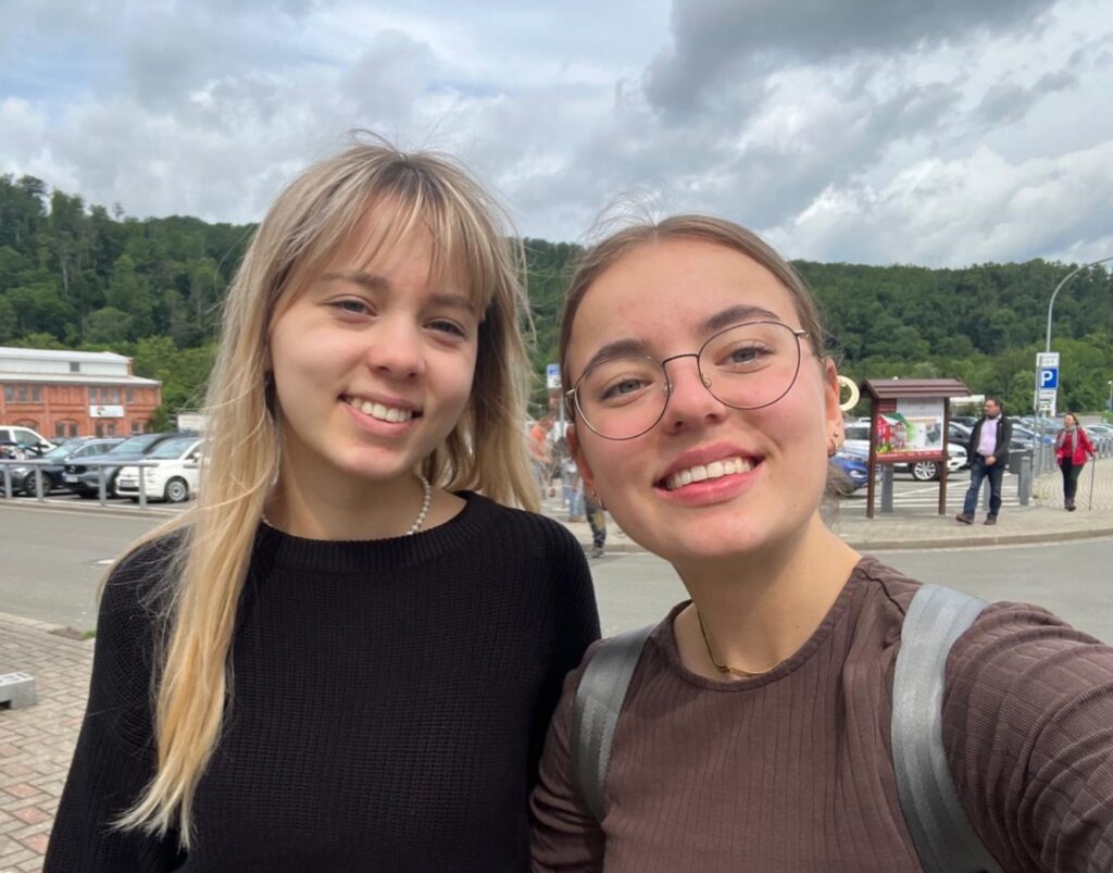 Zwei lächelnde junge Frauen im Vordergrund, eine mit Brille und Rucksack, im Hintergrund Parkplätze, Gebäude und bewaldete Hügel unter bewölktem Himmel.