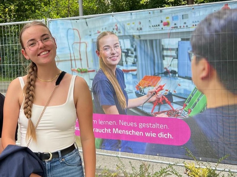 Junge Frau mit Zopf, Brille und weißem Top lächelt vor einem Banner, das sie selbst in einer Werkstatt beim Löten zeigt.