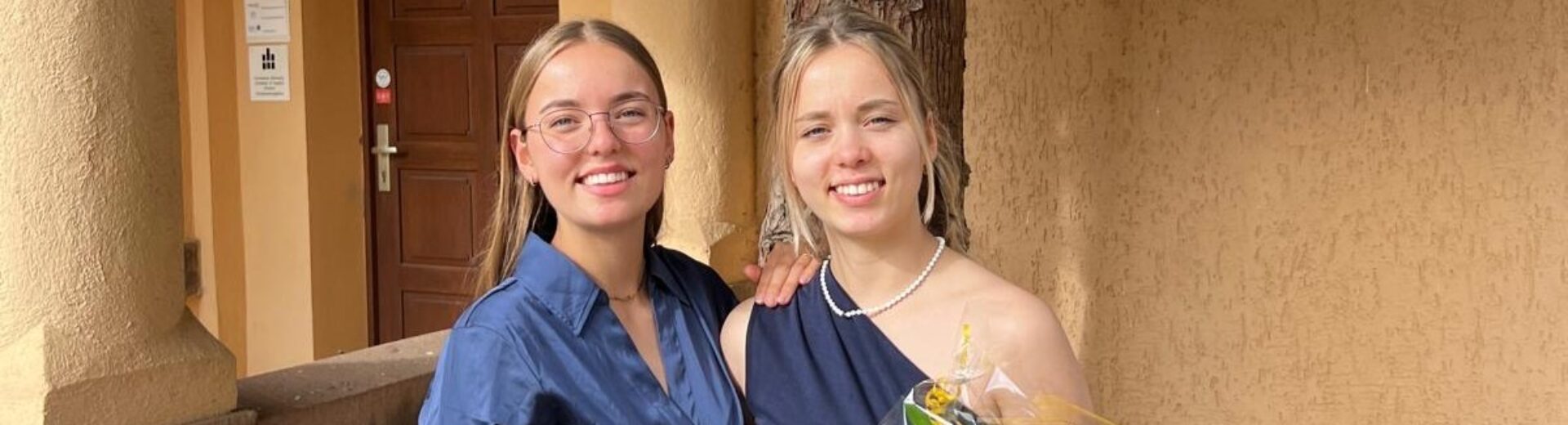 Zwei junge Frauen in eleganter Kleidung vor einem Gebäude; die rechte hält einen Blumenstrauß mit gelber Blume und trägt Perlenkette.