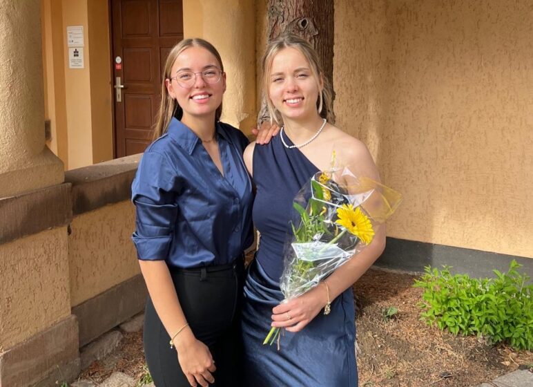 Zwei junge Frauen in eleganter Kleidung vor einem Gebäude; die rechte hält einen Blumenstrauß mit gelber Blume und trägt Perlenkette.