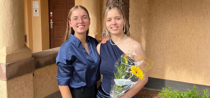 Zwei junge Frauen in eleganter Kleidung vor einem Gebäude; die rechte hält einen Blumenstrauß mit gelber Blume und trägt Perlenkette.
