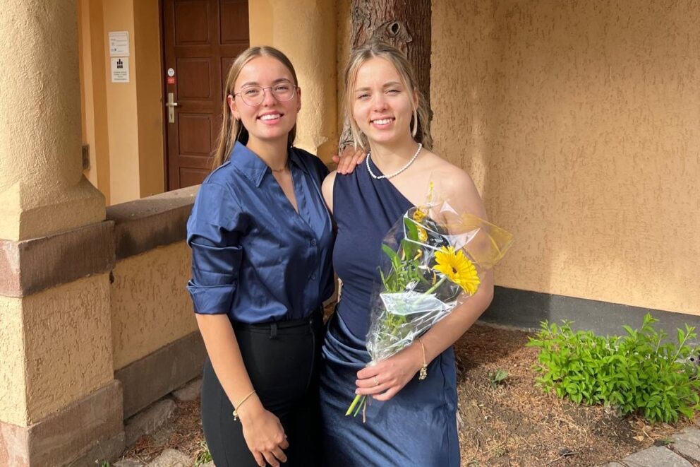 Zwei junge Frauen in eleganter Kleidung vor einem Gebäude; die rechte hält einen Blumenstrauß mit gelber Blume und trägt Perlenkette.