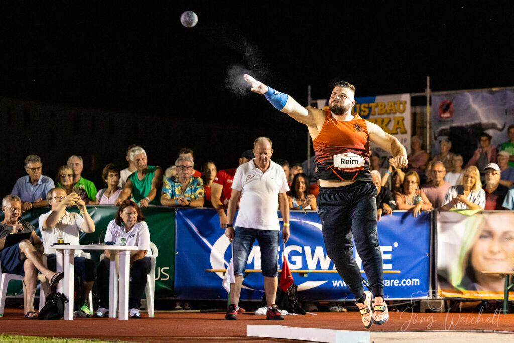 Kugelstoßer in orange Trikot mit Namenstreifen „Reim“ beim Abwurf, Pulverwolke um die Hand und zuschauende Menge im Hintergrund