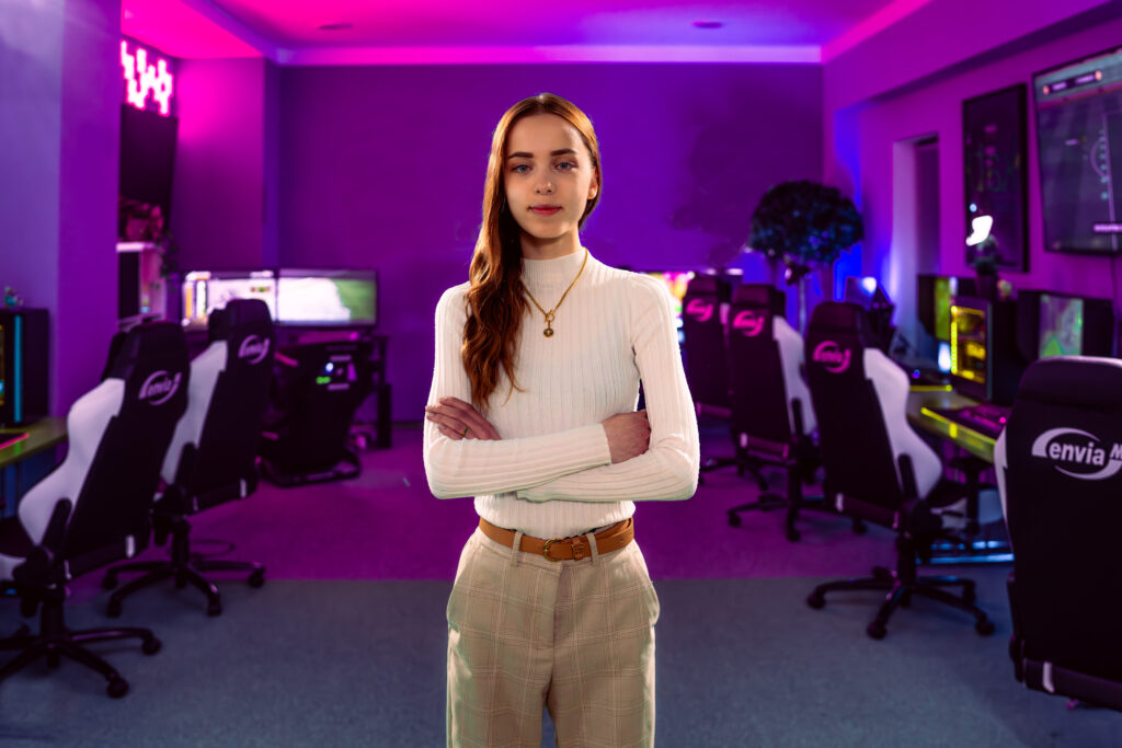 Junge Frau in weißem Rollkragenpullover mit verschränkten Armen vor einer beleuchteten Gaming-Lounge mit PC‑Stühlen und pink-lila LED-Beleuchtung.