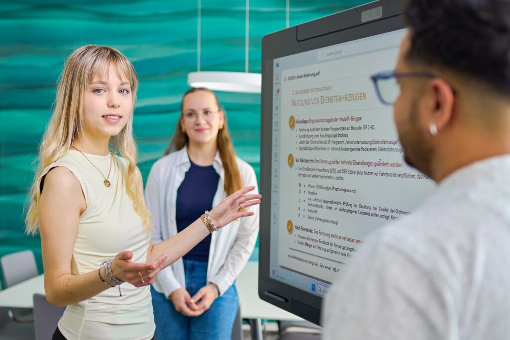 Junge Frau erklärt vor einem großen Bildschirm mit Dokumenten, zwei Kolleg:innen hören im modernen Büro aufmerksam zu.